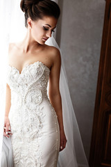 Fototapeta premium Bride in gorgeous wedding dress with crystls and pearls stands before bright window