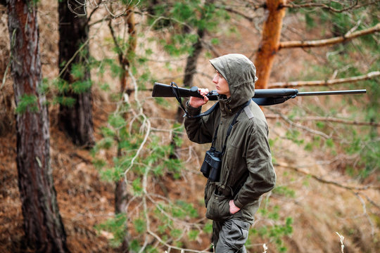male hunter with binoculars ready to hunt, holding gun and walking in forest.