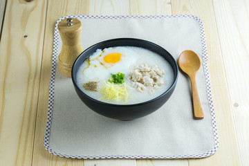 Rice porridge is topping with ginger, spring onion, pepper, chicken, and onsen egg, Breakfast set on wooden table, spoon, pepper grinder. breakfast in Asia call 