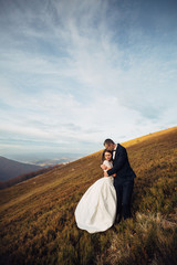 Groom embraces delicate bride standing with her on autumn hill