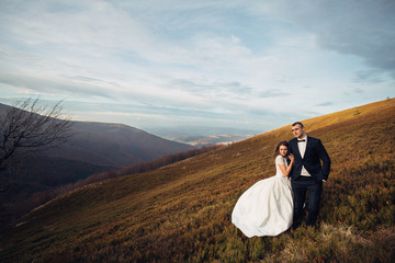Wind blows bride dress while she holds groom's hand tender on autumn hill
