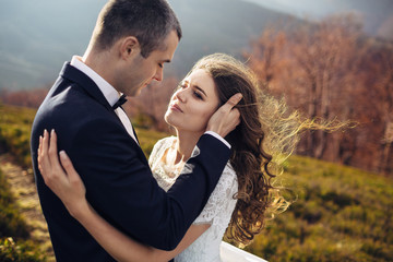 Wind blows bride's hair while groom hugs her tender on the hill