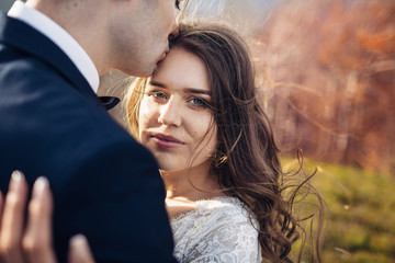 Blue-eyed bride leans to groom's shoulder while he hugs her on the hill