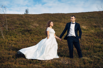 Wind blows bride's gown and groom's jacket while they stand on hill