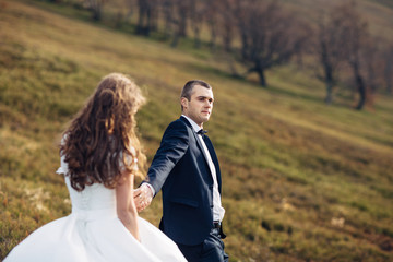 Wind blows bride's gown and groom's jacket while they stand on hill