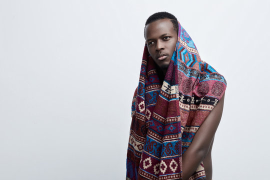 Close up shot of attractive dark-skinned male with clean healthy skin wearing colorful etnic blanket or scaf, looking serious and thoughtful. Pensive hipster young black man posing indoors in studio 