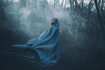 A big, beautiful woman in a blue raincoat, walks in a fog. Background dark forest, bare trees....