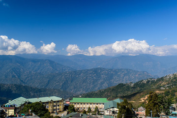 Fototapeta premium A hilltop view of house' roof with mountains and blue sky with clouds from Darjeeling Himalayan railway station on a misty morning.