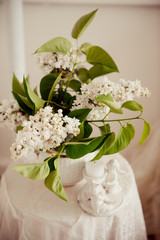 A bouquet of white lilac in a vase on a table. Candlestick with angels. Unburned candle.