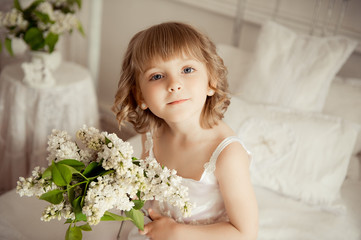 A little girl in a white nightie on a bed with a bouquet of white lilac in her hands.