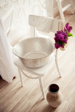 Galvanized basin for washing on a white antique chair. A bouquet of lilac in a basket hangs on the back of a chair.