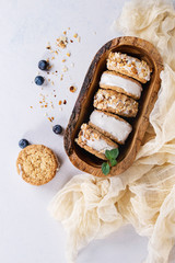 Set of homemade ice cream sandwiches in oat cookies with almond sugar crumbs, blueberries and mint on in olive wooden bowl over gray texture background. Top view with space