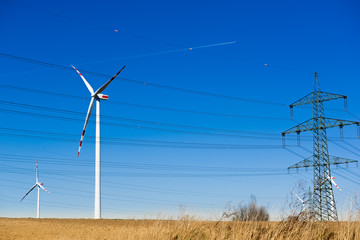 Hochspannungsmast und Windrad vor blauem Himmel mit Feld im Vordergrund