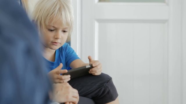 Young attractive mother with adorable cute son sitting in the queue to doctor in clinic, talking and playing games on phone.