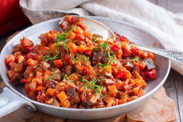 Stewed vegetables (eggplant, peppers, tomatoes) in a frying pan on a wooden table, selective focus,...