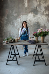female  in gray blouse and jeans make a bouquet over gray background, putting roses in vase, flowers and vase on wood table, workplace