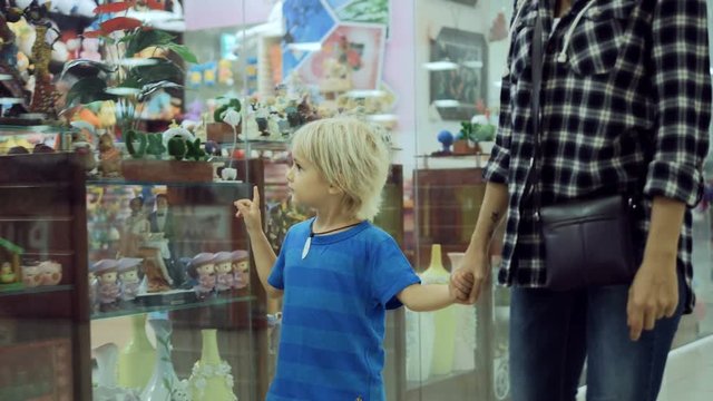 Adorable happy child with young attractive mother walking in big mall along the window displays, looking for and choosing toy. Family shopping in big store.