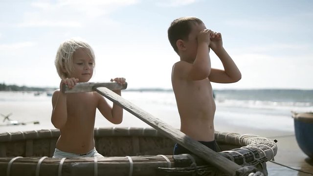 Cute adorable happy children having fun on the sand beach on blue sky and sea background in summer  vacation. Rowing oar sitting in the fishing boat. Playing fishermen, sailors. Vietnam. Slow motion