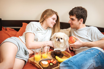 Young couple is lying in bed with dog and having a healthy breakfast.
