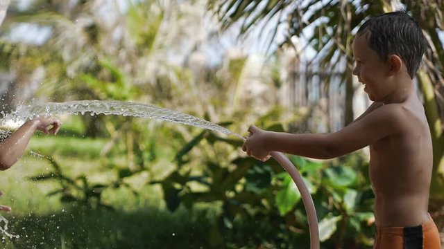 Happy cheerful children playing with water in tropical resort in summer vacation. Cute adorable smiling boys spraying with a garden hose in the back yard. Slow motion