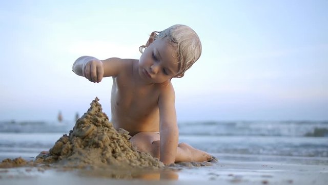 Potrait of cute litlle child making sand castle by himself and broking it on the beach on sea background in sunset. Summer vacation