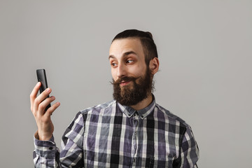 Bearded handsome man is taking selfie by phone in blue squared shirt on grey background