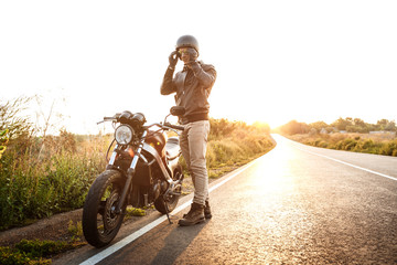 Young handsome man posing near his motorbike at countryside road. Flare sunlight background.