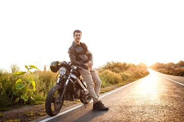 Young handsome man posing near his motorbike at countryside road. Flare sunlight background.