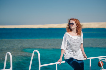 Beautiful ginger woman in sun glasses sits on a white yacht in a sea with clear turquoise water. Relaxation at summer vacation under a sun. Woman looks away.