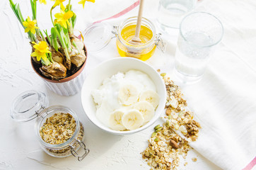 Healthy breakfast fresh granola, muesli in bowl with cereal, nuts, banana fruit, glass of water, bottle, Daffodil flowers. Top view food, Diet, Detox, Clean Eating or Vegetarian.