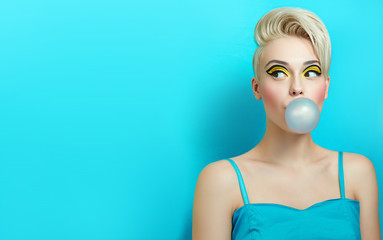 Fashionable girl with a stylish haircut inflates a chewing gum. The girl in the studio on a blue background. The girl's face with bright makeup and yellow with black shadows on the eyes.