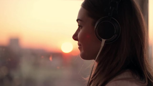 Attractive young woman wears listening to music on the smartphone at city blurred background with sunset. enjoying the tunes in her headphones