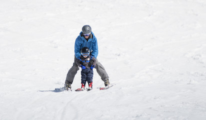 Toddler Boy Skis Downhill with the Help of His Father. Dressed Safely with Helmet and Harness.