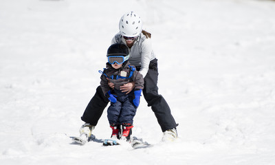 Fototapeta na wymiar Mother Helps Toddler Boy Ski Downhill. Dressed Safely in Helmet and Harness