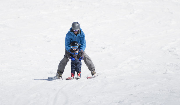 Toddler Boy Skis Downhill with the Help of His Father. Dressed Safely with Helmet and Harness.