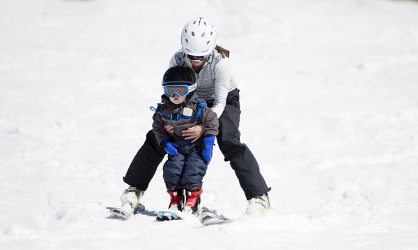 Mother Helps Toddler Boy Ski Downhill. Dressed Safely in Helmet and Harness