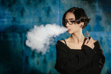 A girl with glasses vaping and releases a cloud of vapor. Model in a black vaper smoke vaporizer on...