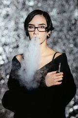 A girl with glasses vaping and releases a cloud of vapor. Model in a black vaper smoke vaporizer