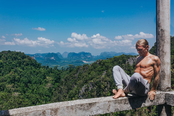 Man stretching body on nature. Young male sitting on narrow balk and stretching on background of picturesque tropical valley.
