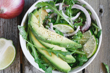 Overhead view on a fresh summer salad from avocado, red onion, arugula, sesame and olive oil in a light bowl on an old wooden table. Healthy food.