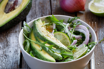 Close-up of a fresh summer salad from avocado, red onion, arugula, sesame and olive oil in a light bowl on an old wooden table. Healthy food.