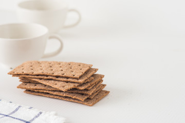 Obraz na płótnie Canvas Stack of brown rye crispy bread (Swedish crackers) with two cups and piece of cloth on white background with space for text. Healthy meal concept