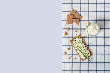 Brown rye crispy bread (Swedish crackers) with spread cottage cheese, decorated with thin green onion, on piece of cloth on light blue background with space for text, top view. Healthy snack concept