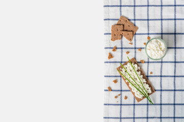 Brown rye crispy bread (Swedish crackers) with spread cottage cheese, decorated with thin green onion, on piece of cloth on white background with space for text, top view. Healthy snack concept