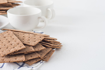 Stack of brown rye crispy bread (Swedish crackers) with two cups and piece of cloth on white background with space for text. Healthy meal concept