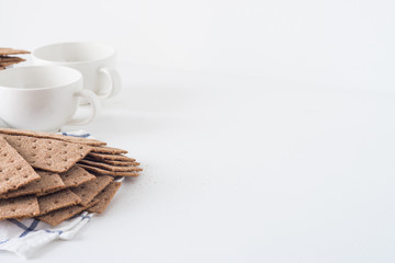 Stack of brown rye crispy bread (Swedish crackers) with two cups and piece of cloth on white background with space for text. Healthy meal concept