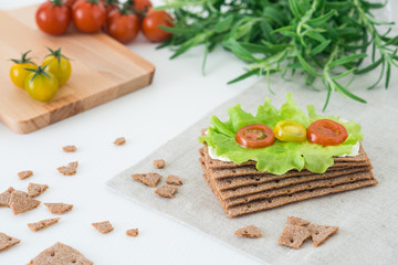 Rye crispy bread (Swedish crackers) with soft cheese, lettuce leave, cherry tomatoes and rosemary on white background. Healthy snack concept