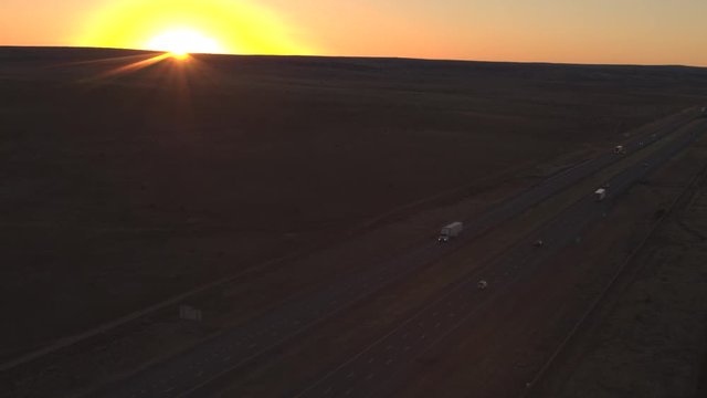AERIAL Flying above the busy heavy traffic interstate highway running through the deserted Great Plains in America. Freight semi trucks transporting goods, cars on journey, people traveling at sunrise