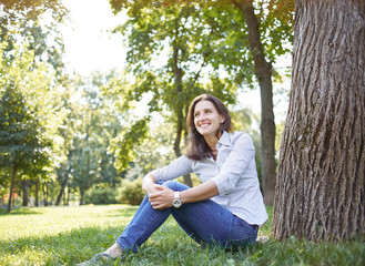 Cute young woman looking like a student sitting in a park on the grass under a tree. On her big headphones, near her is a book