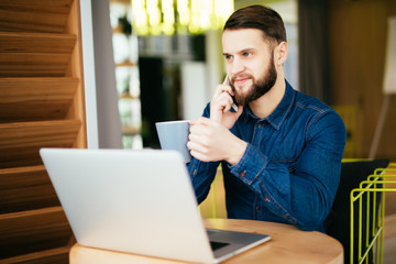 Young handsome man with beard sitting in cafe talking mobile phone, holding cup of coffee and smiling. Laptop on wooden table.